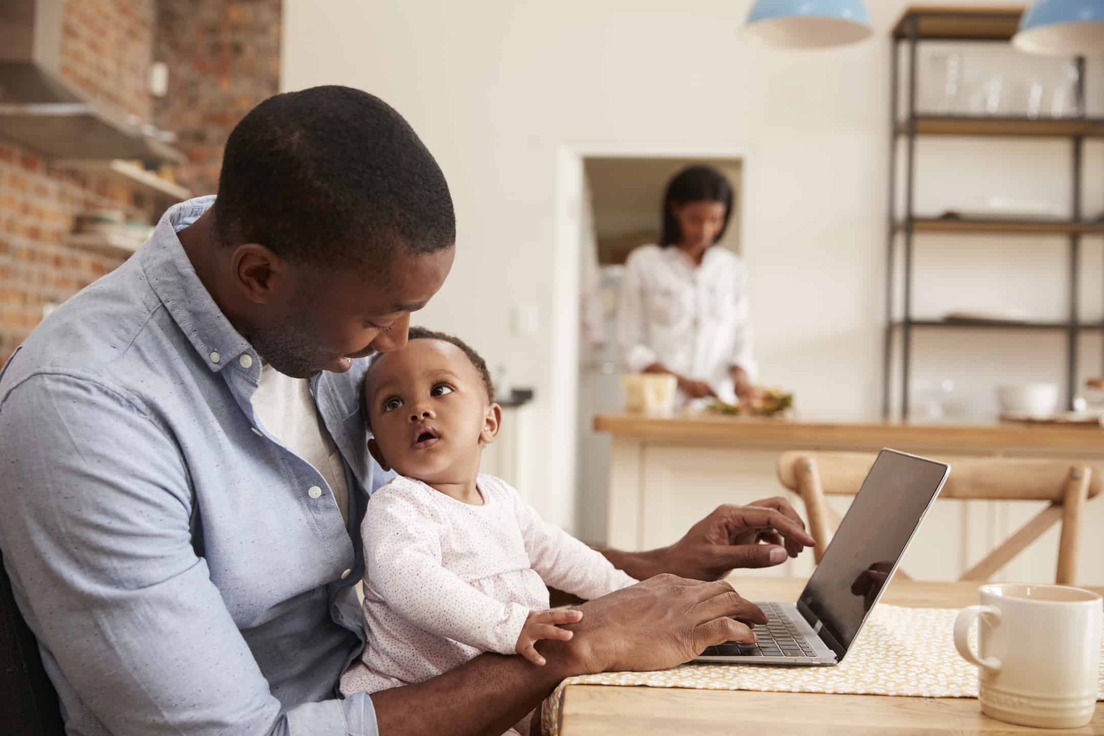 Man Working on Computer at Kitchen Table with Baby Daughter in His Lap