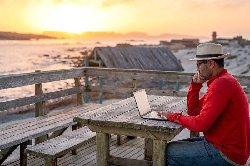 Man on laptop on a deck at sunset by a beach - learn how to start a vacation planning business