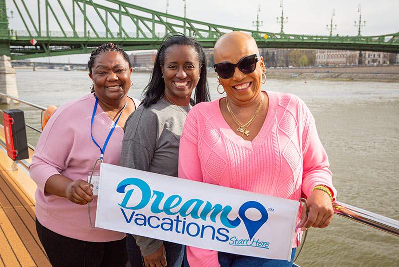 Three women posing in front of a bridge with a Dream Vacations sign