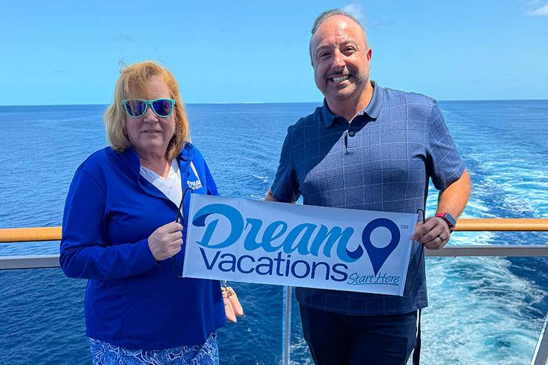 A man and woman on a cruise ship hold a sign that reads "Dream Vacations," smiling at the camera.