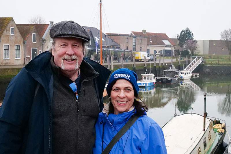 An older couple stands beside a boat in a harbor