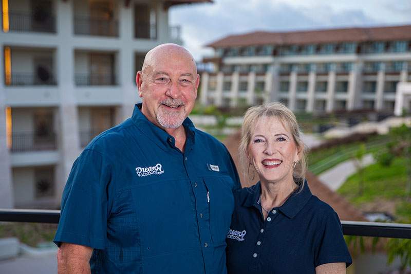 A man and woman stand on a balcony, overlooking a resort in the background.