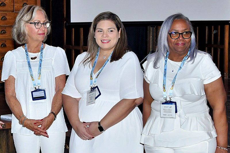 Three women in white attire standing closely together, smiling.